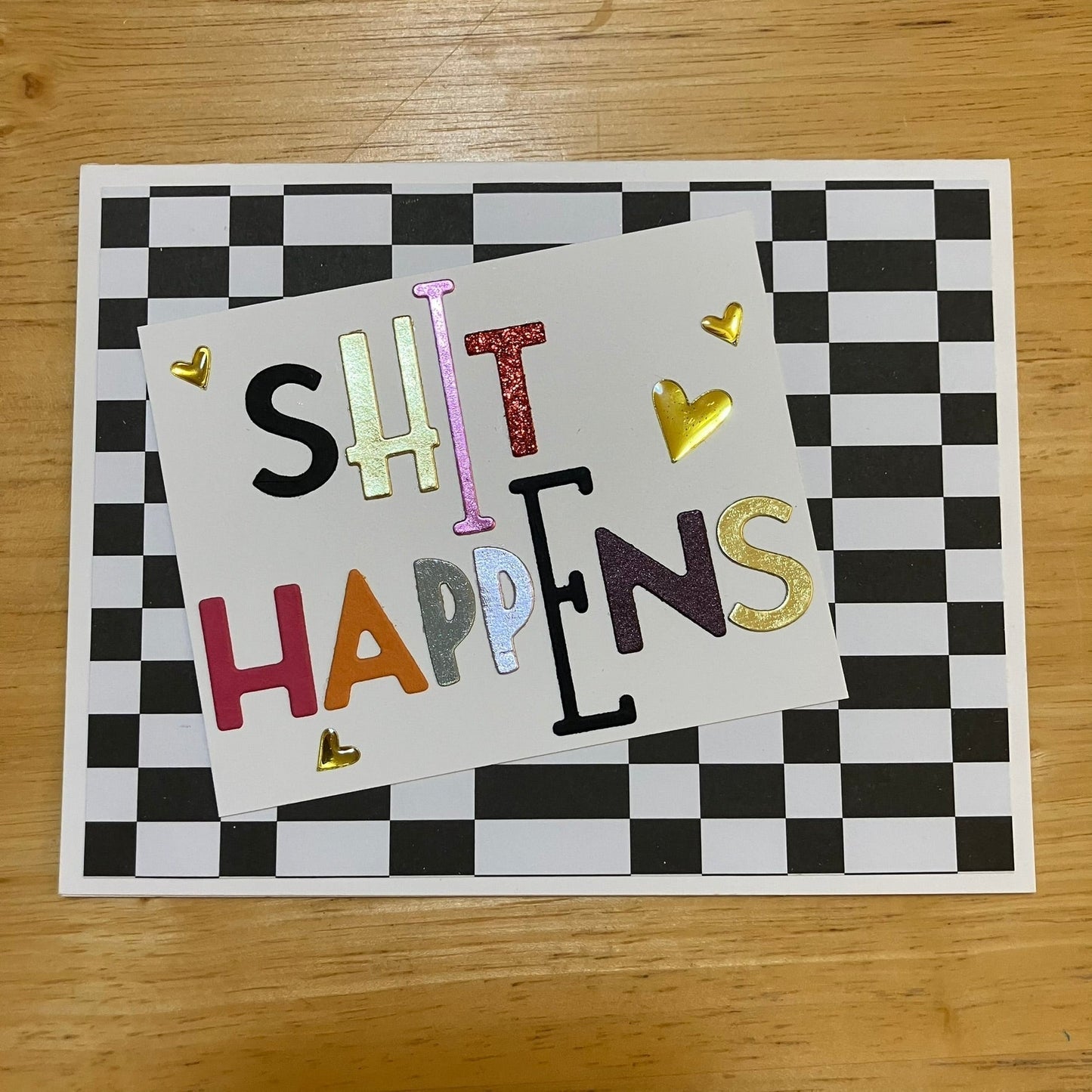 Multicoloured variable sized text text on white angled rectangle with gold heart accents. On black and white checked background. White card, interior black.
