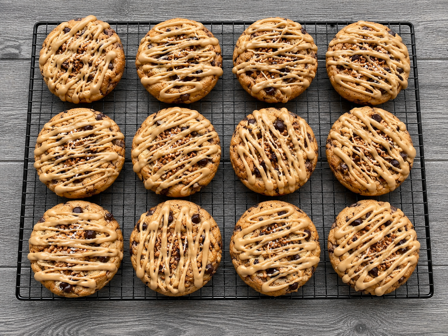 Nine round chocolate chip cookies with crisscrossed glaze drizzle and chopped nuts, arranged on a black wire cooling rack over a gray wood-textured surface.