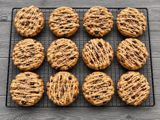 Nine round chocolate chip cookies with crisscrossed glaze drizzle and chopped nuts, arranged on a black wire cooling rack over a gray wood-textured surface.